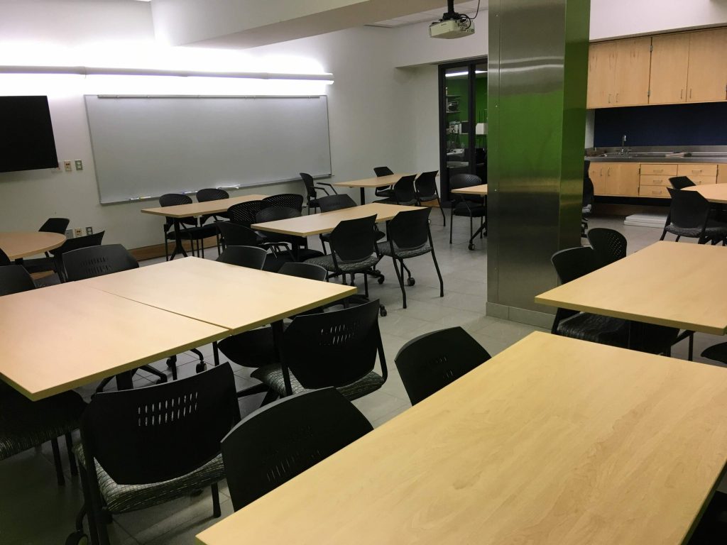 A Classroom, with wooded-topped tables and chairs, a blank whiteboard, a stainless steel column in the middle of the room, along with prep area on the far side
