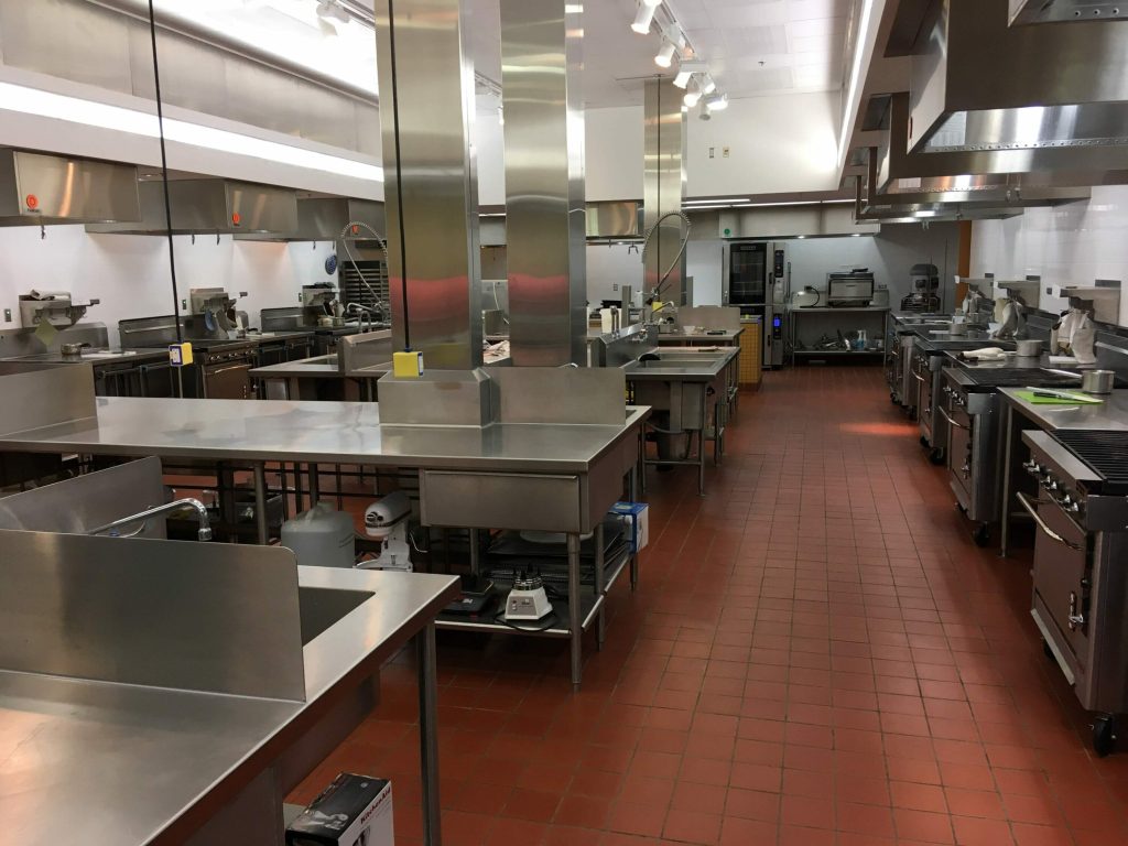 Food Application lab that houses food service equipment, dishwashing stations, and resource areas. It is a red tiled area with stainless steel countertops and appliances.
